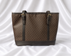 Brown women's monogram-inspired tote bag with dual shoulder straps and gold-tone buckle details.